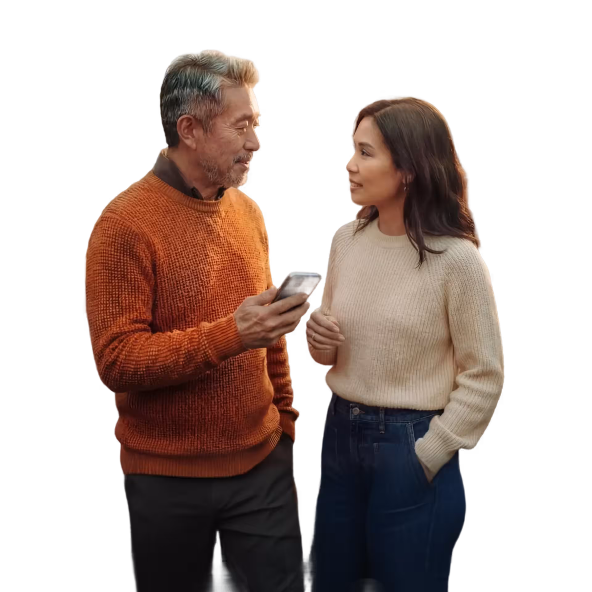 Middle-aged man in orange sweater shows a smartphone to a woman in a cream sweater and jeans as they engage in conversation.