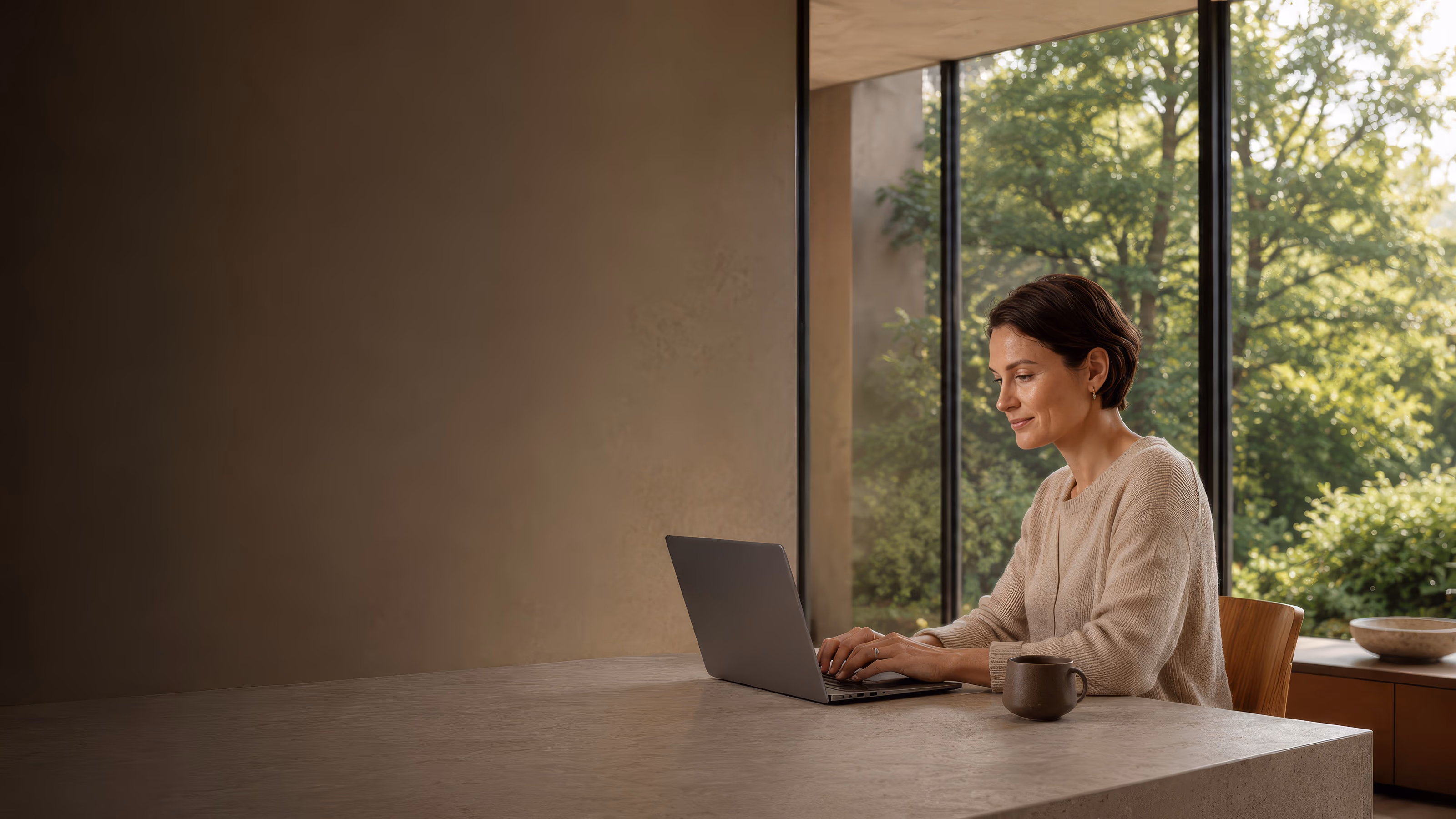 A woman sits at a stone desk with a laptop and a mug, lit by a tall window with green trees behind her.